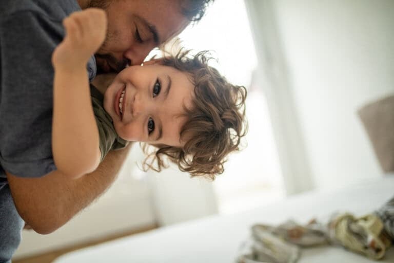 A man is playfully holding a smiling 23-month-old child with curly hair. The child is laughing and facing the camera. The background is softly blurred, showing a bright room and part of a bed, capturing a joyful moment amidst unpredictable sleep schedules.