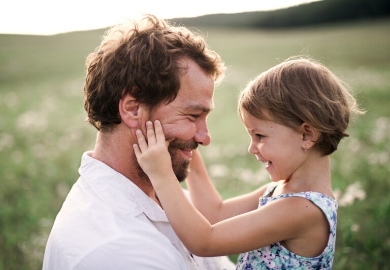 A man with brown hair and a beard, wearing a white shirt, smiles while holding his young daughter in a blue floral dress. They are outdoors in a field with green grass and small white flowers. The girl touches her dad's face and smiles at him.