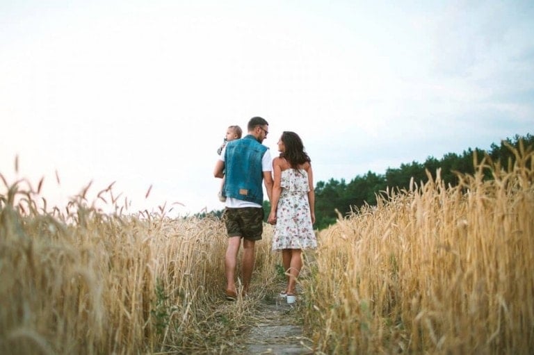 A man, woman, and a child are walking through a golden wheat field. The man carries the child on his shoulders, and they all hold hands, their relationship growing stronger with each step. The sky above is clear with a few clouds, and there are trees visible in the background.