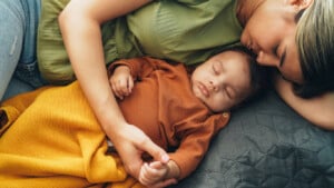 A woman and her 4-month-old baby are lying down together on a quilted surface. The baby, dressed in a brown outfit, is covered with a mustard-colored blanket and is sleeping. The woman, wearing a green top, is resting her head close to the baby's head.