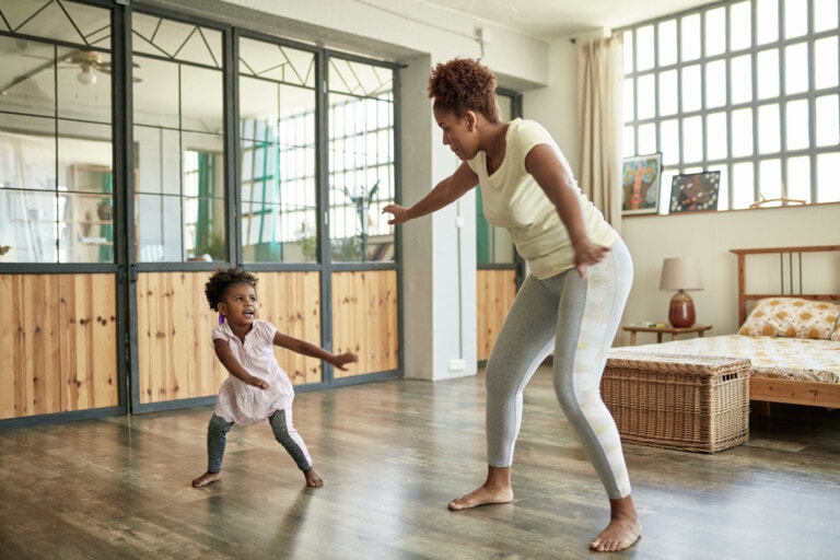 A woman and a young girl are dancing together in a spacious room with large windows. The woman is wearing light-colored workout clothes, and the girl is in a pink dress and gray pants. The room, perfect for activities for kids, has wooden accents and a wicker storage chest.