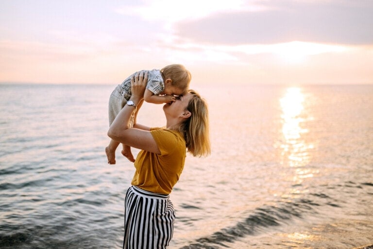 A woman, embodying the essence of being a parent, holds a small child up in the air while standing on a beach at sunset. The woman wears a yellow shirt and striped pants. The child is wearing a patterned shirt and light-colored shorts. The sun is low on the horizon, reflecting off the water.