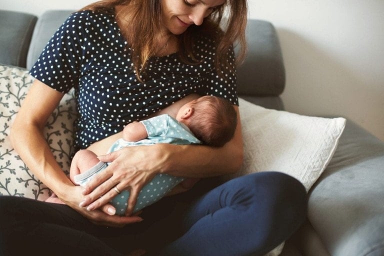 A woman is seated on a couch, breastfeeding an infant with ease, thanks to her breastfeeding essentials. She is wearing a polka-dotted shirt and holding the baby, who is dressed in a light blue onesie, close to her chest. Patterned pillows add comfort in the background.