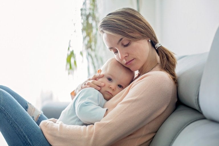 A woman with long hair sits on a couch, holding a baby close. The baby rests its head on the woman's chest, looking towards the camera. The woman has her eyes closed and appears to be comforting the baby amidst her struggle with post-weaning depression. They are in a bright, softly lit room.