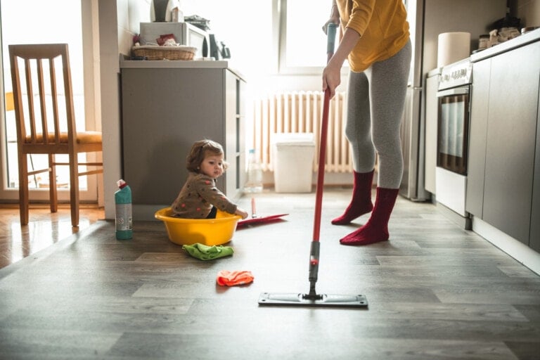 A person mops the floor in a kitchen while a child sits in a plastic tub on the floor. The kitchen has grey cabinets and a wooden chair. Various cleaning supplies, including a bottle of cleaner with potential toxins and cloths, are on the floor nearby.