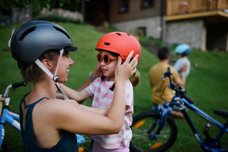 An adult adjusts the orange helmet of a child wearing sunglasses and a striped shirt—one of the signs you're a good parent. In the background, another adult and child with a bicycle sit on the grass.