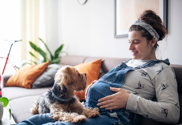 A person wearing overalls and a long-sleeved shirt sits on a couch, resting their hands on their pregnant belly—one of the many things pregnant women do alone to cherish these moments. A small dog sits next to them, looking up at the person. There are orange cushions, a bicycle, and a houseplant in the background.