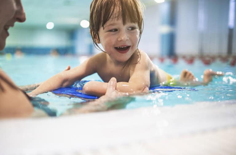 A child with wet hair is lying on a blue kickboard in a swimming pool, supported and guided by an adult during swim lessons. The indoor pool area is well-lit and features red lane markers in the background.