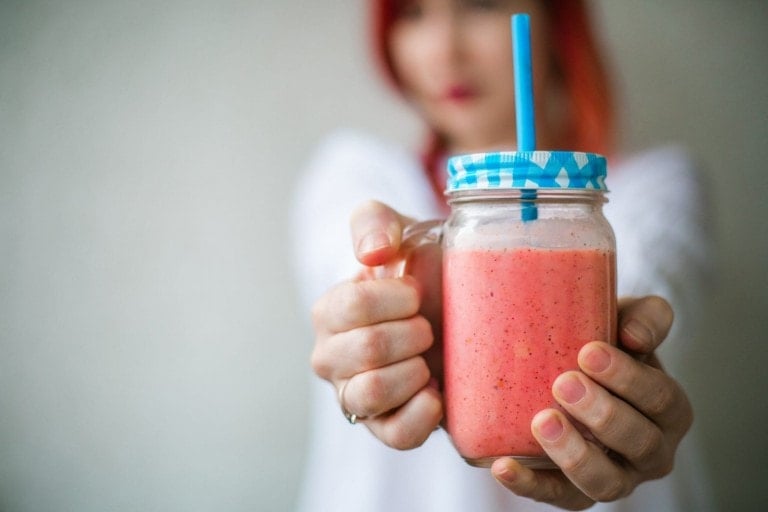 A person with red hair holds out a mason jar with a blue checkered lid and a blue straw. The jar contains a pink lactation smoothie. The background is plain and out-of-focus.