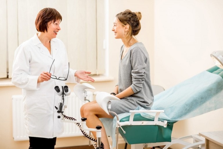 A doctor in a white coat is talking to a seated patient in a medical examination room. During the 6-week postpartum checkup, the patient sits on an examination chair covered with a blue sheet. The doctor, holding a pair of eyeglasses and gesturing with her hand, offers reassuring advice.