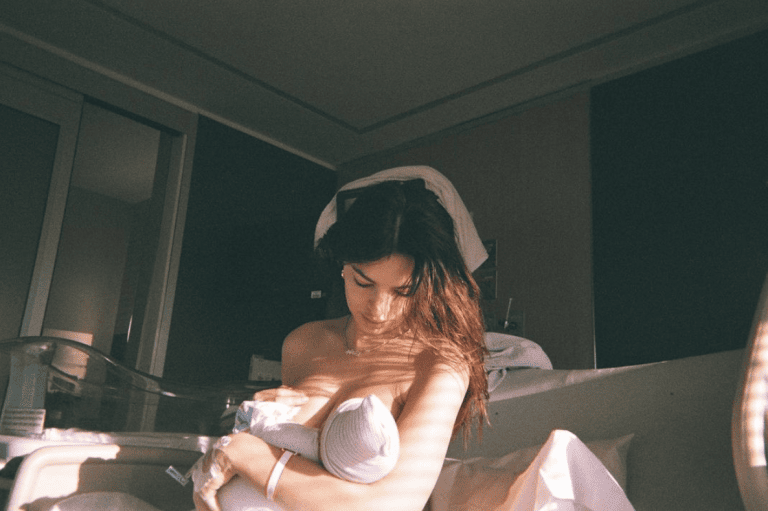 A woman sits on a hospital bed, breastfeeding a swaddled newborn baby. The room is softly lit by natural light streaming in from the window. Reminiscent of Emily Ratajkowski's serene maternity photos, she has long, dark hair and gazes down at the baby in her arms.