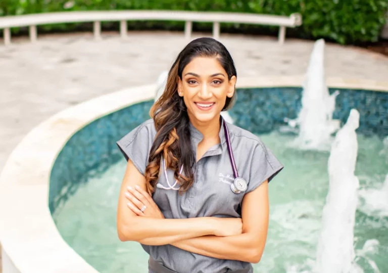A person with long, wavy hair, wearing gray medical scrubs with a stethoscope around their neck, stands in front of a fountain with arms crossed. Surrounded by lush greenery and enjoying the summer warmth, they exude an aura of dedication to health and wellness.