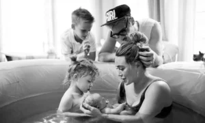 A woman is in a birthing pool, holding a newborn baby while a young child looks on. A man stands beside the pool, gazing at the newborn, with another child standing near him. The scene, reminiscent of celebrities who had home births, highlights the intimate and serene environment of welcoming new life at home.