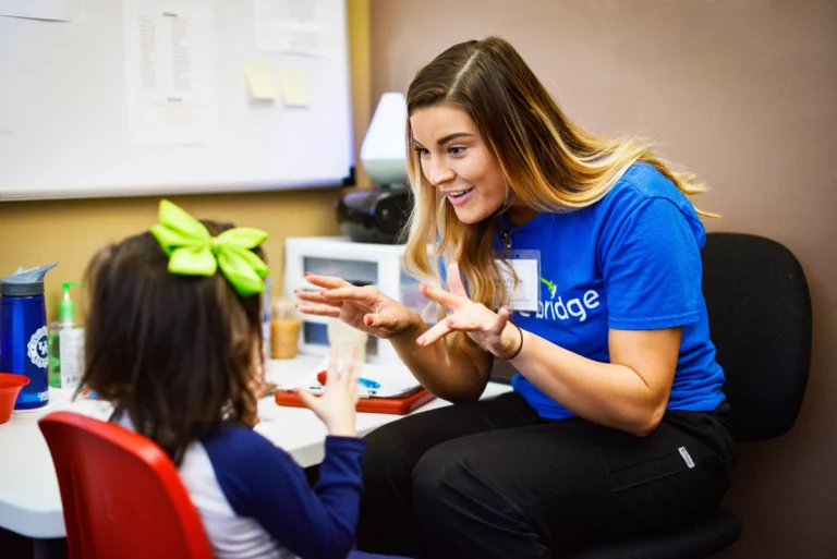 A woman with long hair, wearing a blue shirt, is sitting at a desk and engaging in a lively conversation with a young girl who is sitting on a red chair. The girl, adorned with a large green bow in her hair, shows no autism signs or symptoms. The background includes a whiteboard and desk items.